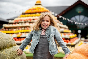 Herbst auf dem Krewelshof Ein lächelndes Mädchen posiert vor einer großen, bunten Pyramide aus Kürbissen verschiedener Farben.