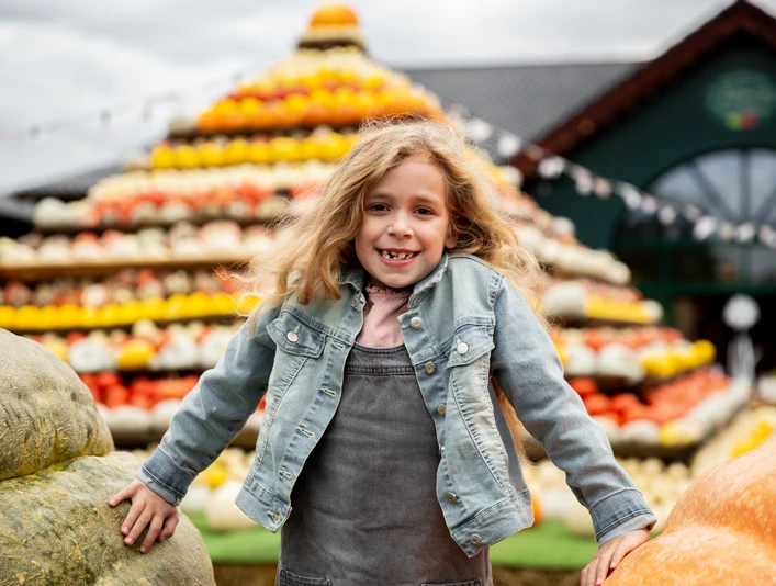 Herbst auf dem Krewelshof Ein lächelndes Mädchen posiert vor einer großen, bunten Pyramide aus Kürbissen verschiedener Farben.
