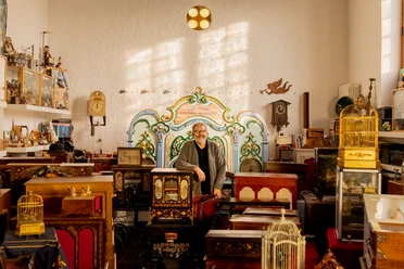 Drehorgelmuseum Ein Mann steht in einem Raum voller antiker Drehorgeln und Uhren, umgeben von nostalgischem Dekor.