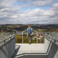 Skywalk Morsbach Frau mit blauem Rucksack genießt von Aussichtsplattform einen weiten Blick über bewaldete Hügel.