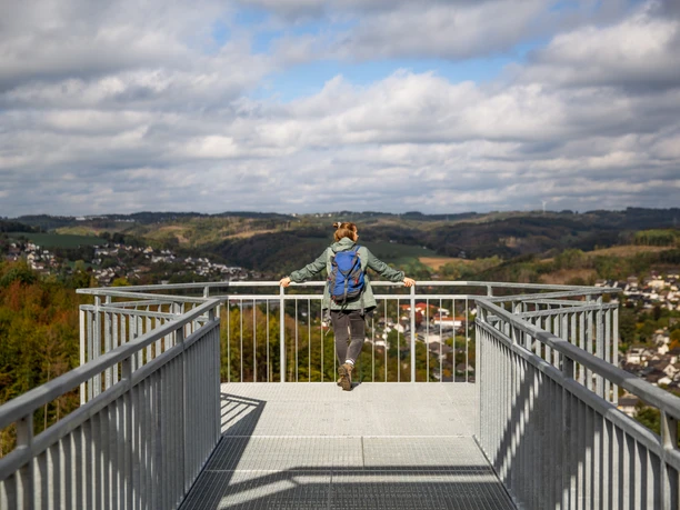 Skywalk Morsbach Frau mit blauem Rucksack genießt von Aussichtsplattform einen weiten Blick über bewaldete Hügel.