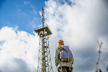 Aussichtsturm Hohe Hardt Person mit Rucksack betrachtet bei bewölktem Himmel einen hohen Sendemast aus Metall.