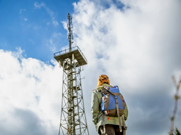 Aussichtsturm Hohe Hardt Person mit Rucksack betrachtet bei bewölktem Himmel einen hohen Sendemast aus Metall.