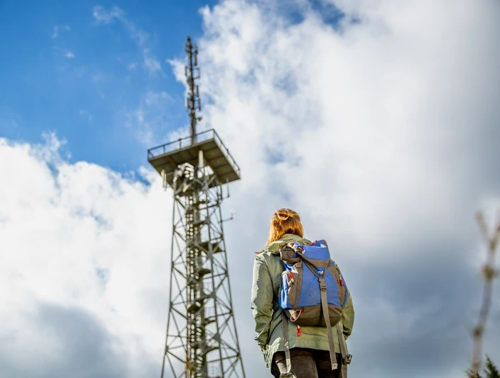 Aussichtsturm Hohe Hardt Person mit Rucksack betrachtet bei bewölktem Himmel einen hohen Sendemast aus Metall.