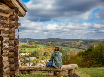 Pause am Aussichtsturm Hohe Hardt <p>Person sitzt auf Bank vor Holzhütte mit Blick auf eine hügelige, bewaldete Landschaft unter bewölktem Himmel.</p>