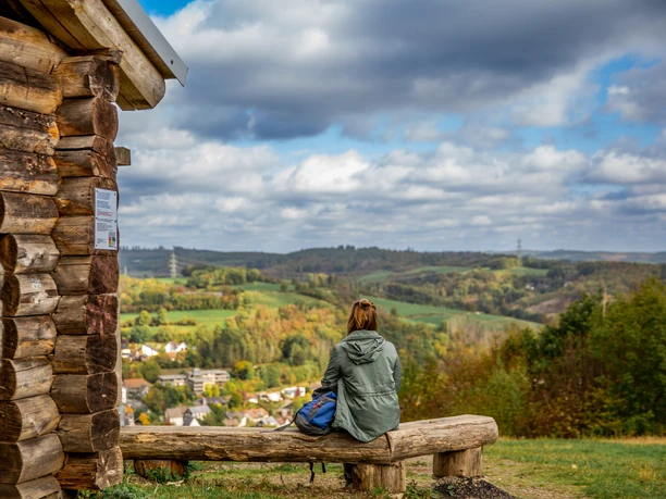 Pause am Aussichtsturm Hohe Hardt <p>Person sitzt auf Bank vor Holzhütte mit Blick auf eine hügelige, bewaldete Landschaft unter bewölktem Himmel.</p>
