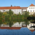 Landesvolkshochschule Hardehausen Die Landesvolkshochschule Hardehausen spiegelt sich in einem ruhigen See, umgeben von grüner Natur.
