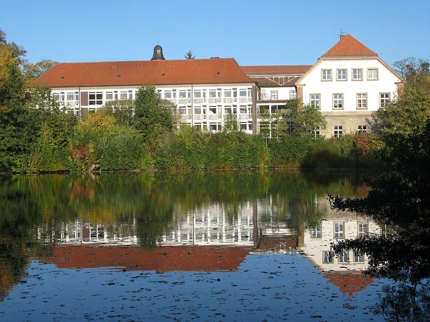 Landesvolkshochschule Hardehausen Die Landesvolkshochschule Hardehausen spiegelt sich in einem ruhigen See, umgeben von grüner Natur.