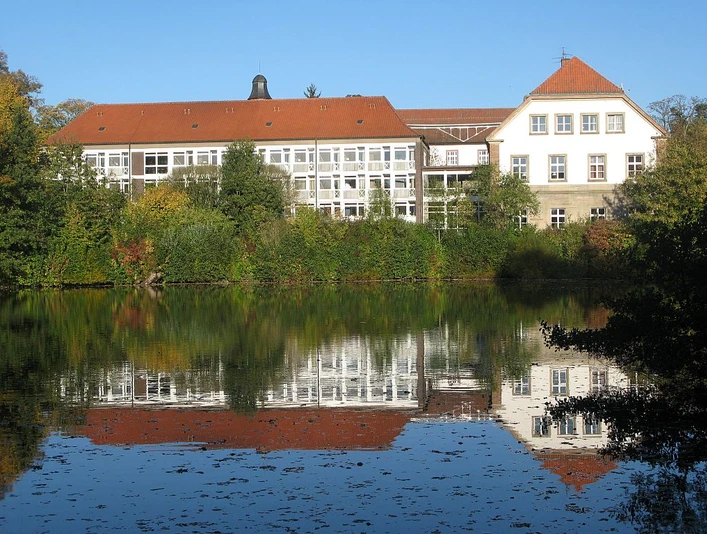 Landesvolkshochschule Hardehausen Die Landesvolkshochschule Hardehausen spiegelt sich in einem ruhigen See, umgeben von grüner Natur.