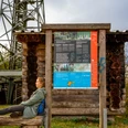 Infotafel am Baumweg Frau sitzt entspannt vor einem hölzernen Wegweiser mit Informationstafel, im Hintergrund ein Turm.