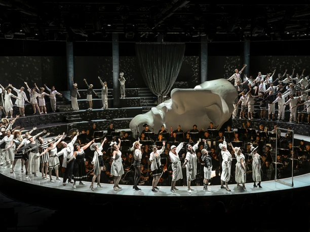 Cologne Opera in the StaatenHaus A stage full of gray and white costumes, a choir behind it; a monumental head sculpture dominates.
