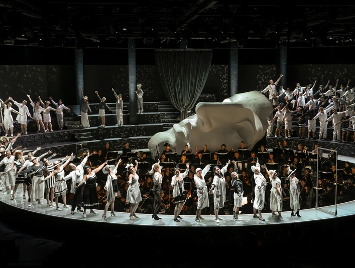 Cologne Opera in the StaatenHaus Eine Bühne voller grau-weißer Kostüme, ein Chor dahinter; eine monumentale Kopfskulptur dominiert.A stage full of gray and white costumes, a choir behind it; a monumental head sculpture dominates.