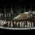 Oper Köln im StaatenHaus Eine Bühne voller grau-weißer Kostüme, ein Chor dahinter; eine monumentale Kopfskulptur dominiert.A stage full of gray and white costumes, a choir behind it; a monumental head sculpture dominates.
