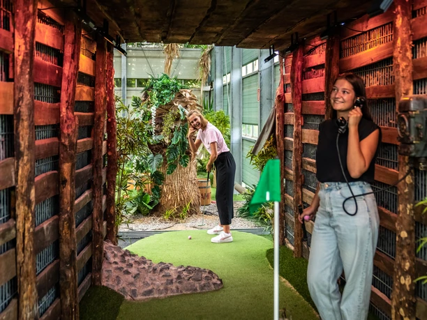 Adventuregolf Zwei Frauen spielen Minigolf in einem tropisch gestalteten Indoor-Parcours aus Holz und Pflanzen.