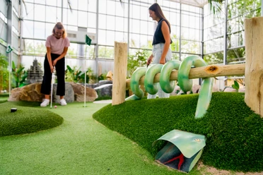Adventuregolf Zwei Frauen spielen Minigolf in einer Indooranlage mit natürlichen Elementen und viel Grün.