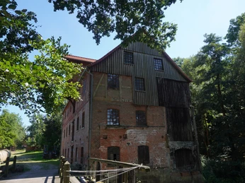 Alte Backsteinmühle in Eitze, umgeben von Bäumen, unter blauem Himmel, mit Holzbrücke im Vordergrund.