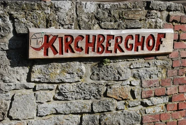 Kirchberghof Herlinghausen Holzschild mit roter Aufschrift "Kirchberghof" hängt an einer groben Steinmauer.
