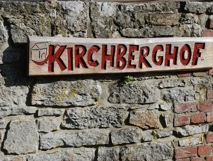 Kirchberghof Herlinghausen Holzschild mit roter Aufschrift "Kirchberghof" hängt an einer groben Steinmauer.