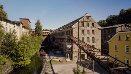 Wülfing Museum Historische Fabrikgebäude umgeben von üppigem Grün und einem ruhig fließenden Fluss bei Sonnenschein.