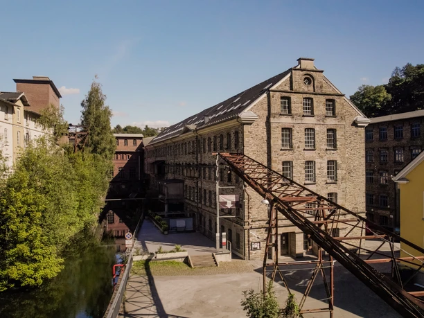Wülfing Museum Historische Fabrikgebäude umgeben von üppigem Grün und einem ruhig fließenden Fluss bei Sonnenschein.