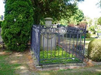 Ein altes Grabmal aus Stein, umgeben von einem kunstvoll verzierten Metallzaun, eingebettet in Grün.