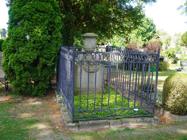Goldmann Grab Ein altes Grabmal aus Stein, umgeben von einem kunstvoll verzierten Metallzaun, eingebettet in Grün.