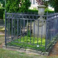 Grabstätte mit schmiedeeisernem Gitter, umgeben von grüner Vegetation, Parkbank in der Nähe.