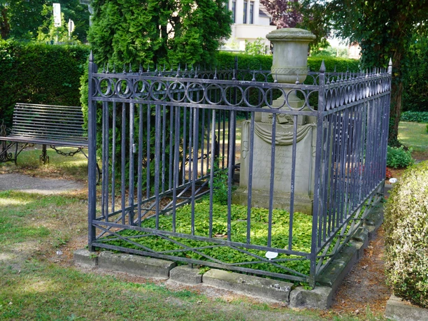 Grabstätte mit schmiedeeisernem Gitter, umgeben von grüner Vegetation, Parkbank in der Nähe.