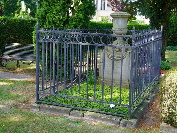 Grabstätte mit schmiedeeisernem Gitter, umgeben von grüner Vegetation, Parkbank in der Nähe.