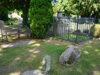 Grabmal mit schmiedeeisernem Zaun, flankiert von hohen Bäumen und einer Bank auf sonnigem Friedhof.