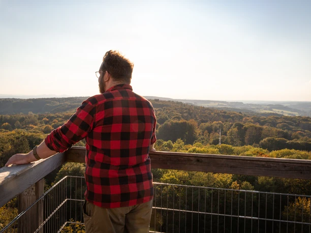 Blick vom Baumwipfelpfad Ein Mann in rot-schwarzem Flanell schaut von einem Aussichtsturm auf einen bewaldeten Hügel.