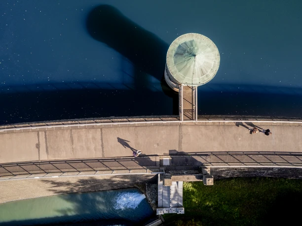 Neyetalsperre Draufsicht auf einen Staudamm mit Rundturm, zwei Spaziergänger auf dem Weg, blauer See im Hintergrund.