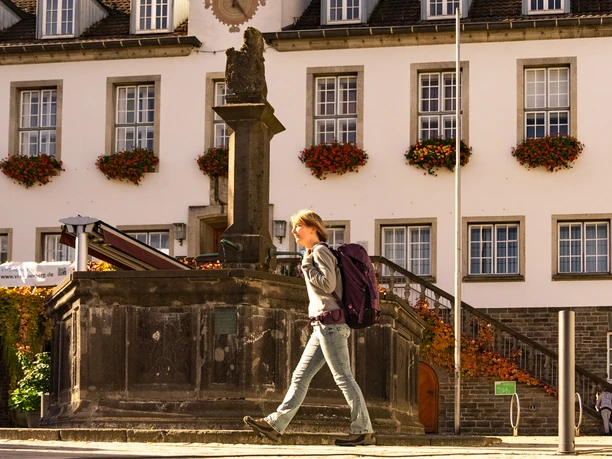 Marktplatz Eine Frau mit Rucksack geht an einem historischen Brunnen vor einem Gebäude mit Blumenkästen vorbei.