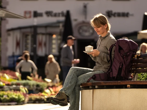 Pause auf dem Marktplatz Eine Frau sitzt auf einer Bank im Freien, hält eine Tasse Kaffee und genießt die Umgebung.