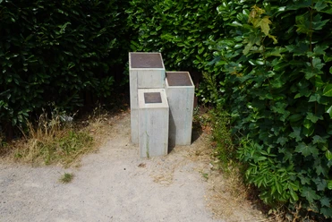 Grau-verwitterte Skulptur zu Ehren von John Lennon, eingebettet in grünen Büschen an einem sandigen Weg.