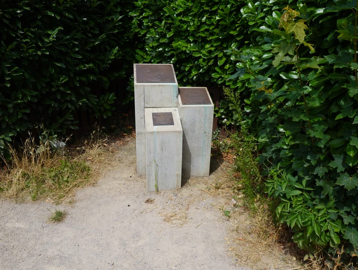 Grau-verwitterte Skulptur zu Ehren von John Lennon, eingebettet in grünen Büschen an einem sandigen Weg.