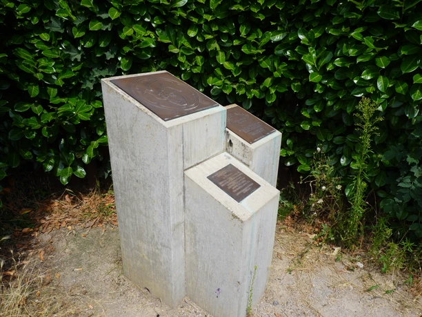A three-part monument made of concrete blocks with embedded metal plates, set against a lush green backdrop.