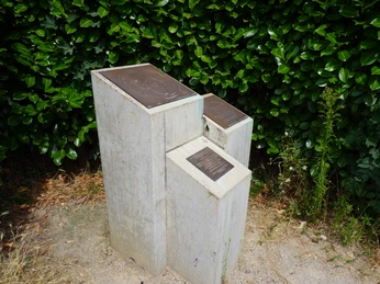 John Lennon-Denkmal Verden Dreiteiliges Denkmal aus Betonquadern mit eingelassenen Metallplatten vor üppig grünem Hintergrund.