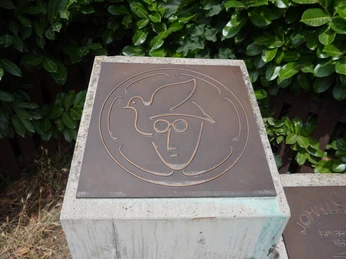 Kupfertafel mit John Lennons stilisiertem Gesicht und Friedenstaube bei dichtem Blattwerk im Hintergrund.