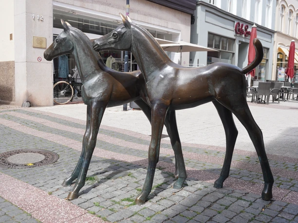 Bronzeskulptur zweier junger Fohlen auf gepflastertem Platz in Verden, umgeben von Geschäften.
