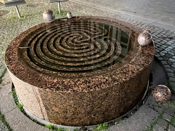Runder Brunnen aus Granit mit spiralförmigem Wasserlauf, umgeben von fünf Kugelskulpturen auf Pflastern.
