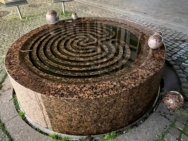 Gezeitenbrunnen Verden Runder Brunnen aus Granit mit spiralförmigem Wasserlauf, umgeben von fünf Kugelskulpturen auf Pflastern.