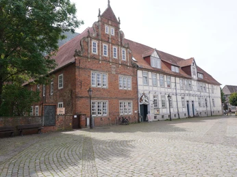 Alter Backsteinbau mit Treppengiebel und Fachwerk, umgeben von Kopfsteinpflaster und Bäumen.