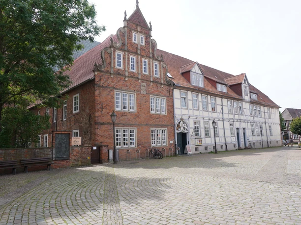 An old brick building with a stepped gable and half-timbering, surrounded by cobblestones and trees.