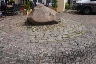 Lugenstein Verden Großer, glattpolierter Stein auf einem Pflasterkreismuster, umgeben von kleinen Sitzgelegenheiten.