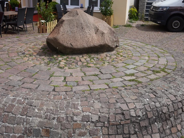Lugenstein Verden Großer, glattpolierter Stein auf einem Pflasterkreismuster, umgeben von kleinen Sitzgelegenheiten.