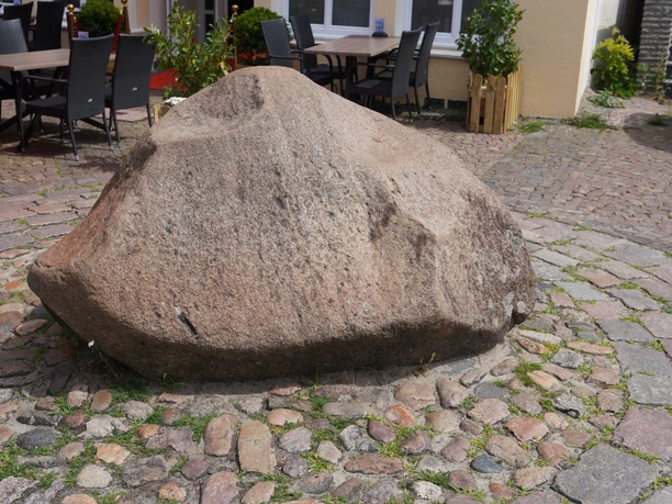 A large, round stone boulder in a paved square, surrounded by chairs and plants.
