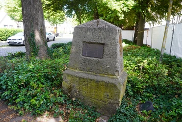 Gedenkstein von Büschen umgeben, mit Plakette darauf, in einem von Bäumen beschatteten Gebiet.