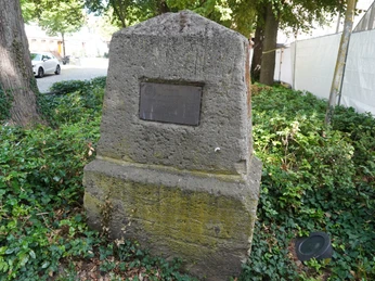Steinmonument mit eingelassener Tafel in einer grünen Parkumgebung, dahinter Bäume und Fahrzeuge.