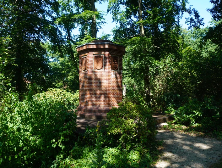 Steinsäule mit Wappen im Schatten grüner Bäume, umgeben von dichtem Laub im Bürgerpark Verden.